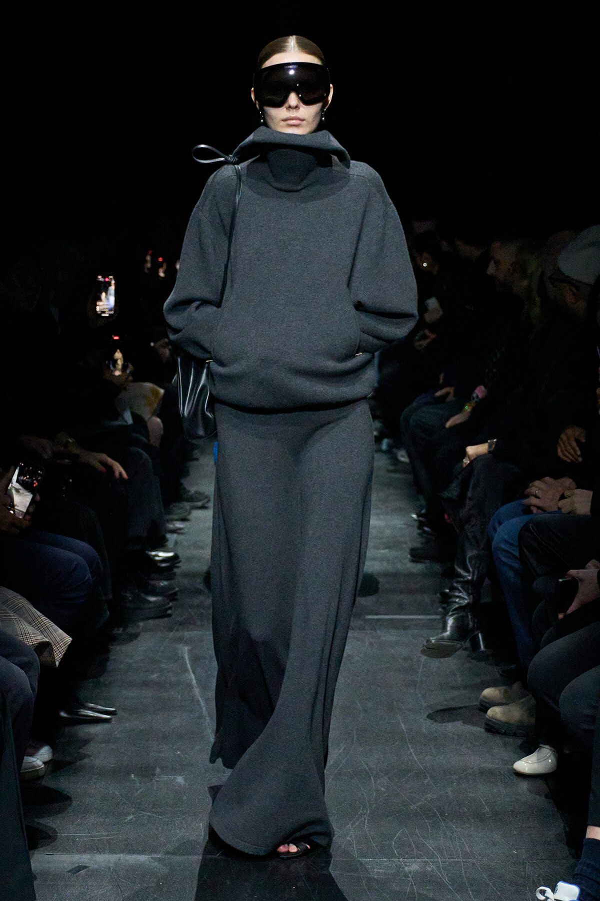 Model on a runway wearing a dark gray oversized knit top with a high collar and matching maxi skirt, plus large black visor sunglasses.