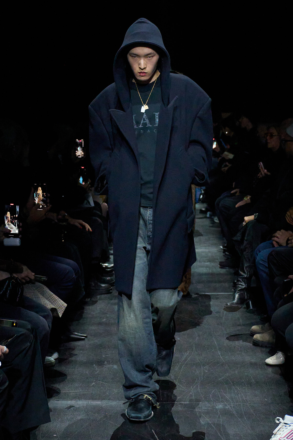 Male model walks a dim runway in a navy oversized coat over a dark hoodie and jeans, surrounded by seated spectators.