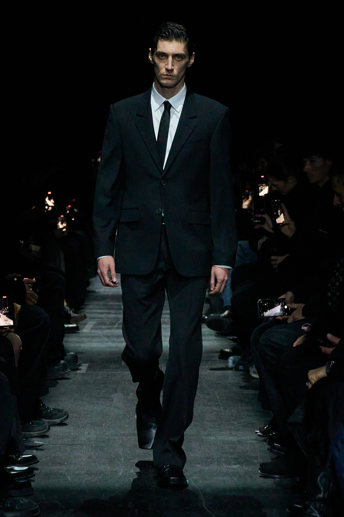 Male model in a black suit and white shirt walking a runway during a fashion show.