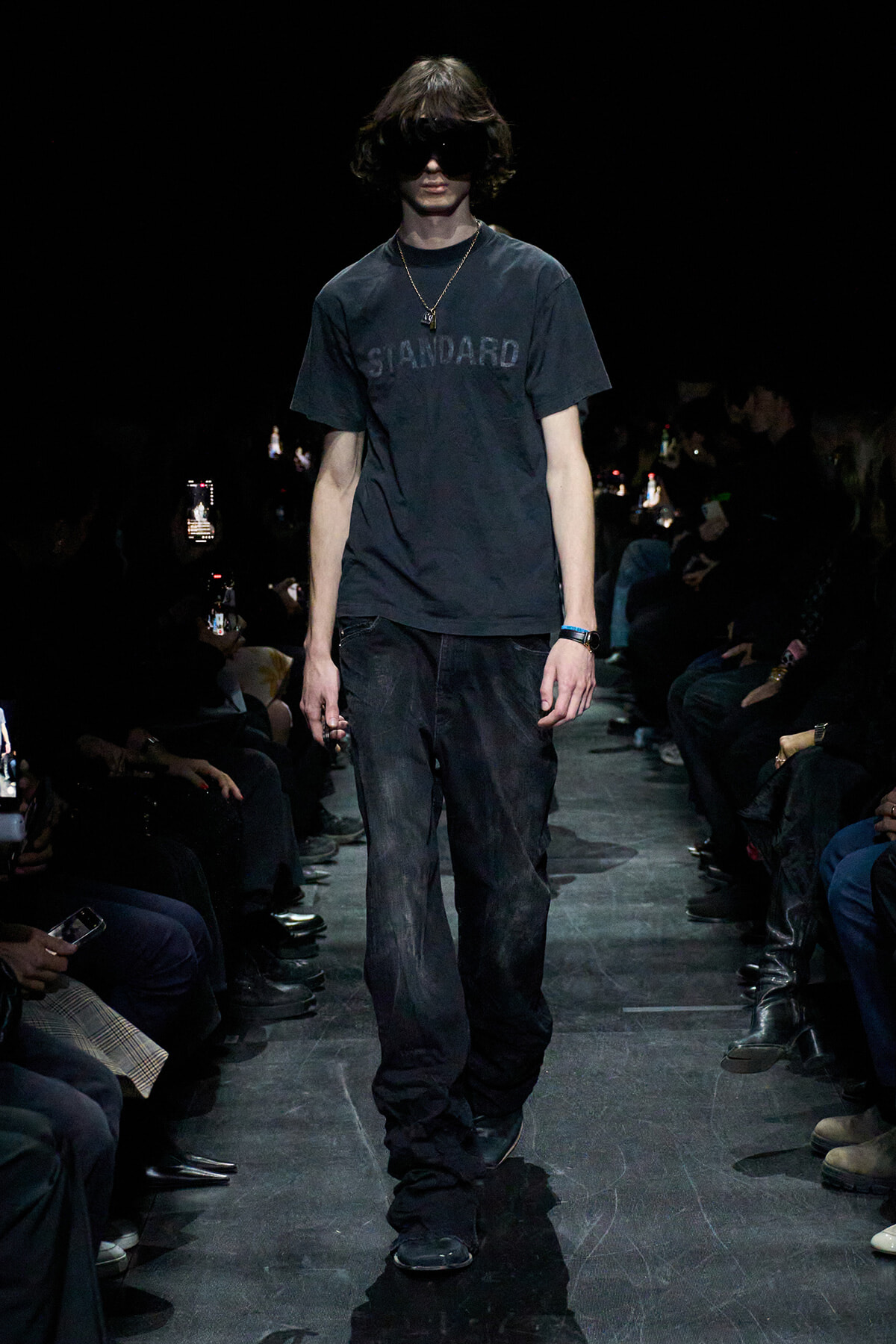 Male model walking a dim runway in a dark 'STANDARD' T‑shirt, black jeans, and oversized sunglasses, accessorized with a necklace.