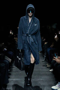 Model walks a fashion runway in a navy hooded robe with white piping, belt tied at the waist, wearing futuristic sunglasses and carrying a black bag on the left.