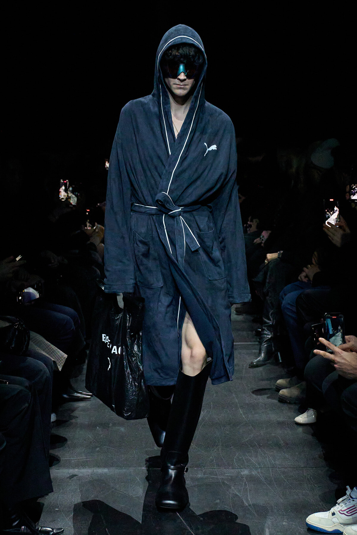 Model walks a fashion runway in a navy hooded robe with white piping, belt tied at the waist, wearing futuristic sunglasses and carrying a black bag on the left.