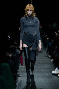Model struts down a dark runway in a fitted dark grey top, black wrapped mini skirt, and tall black boots, with orange hair and a pendant necklace.