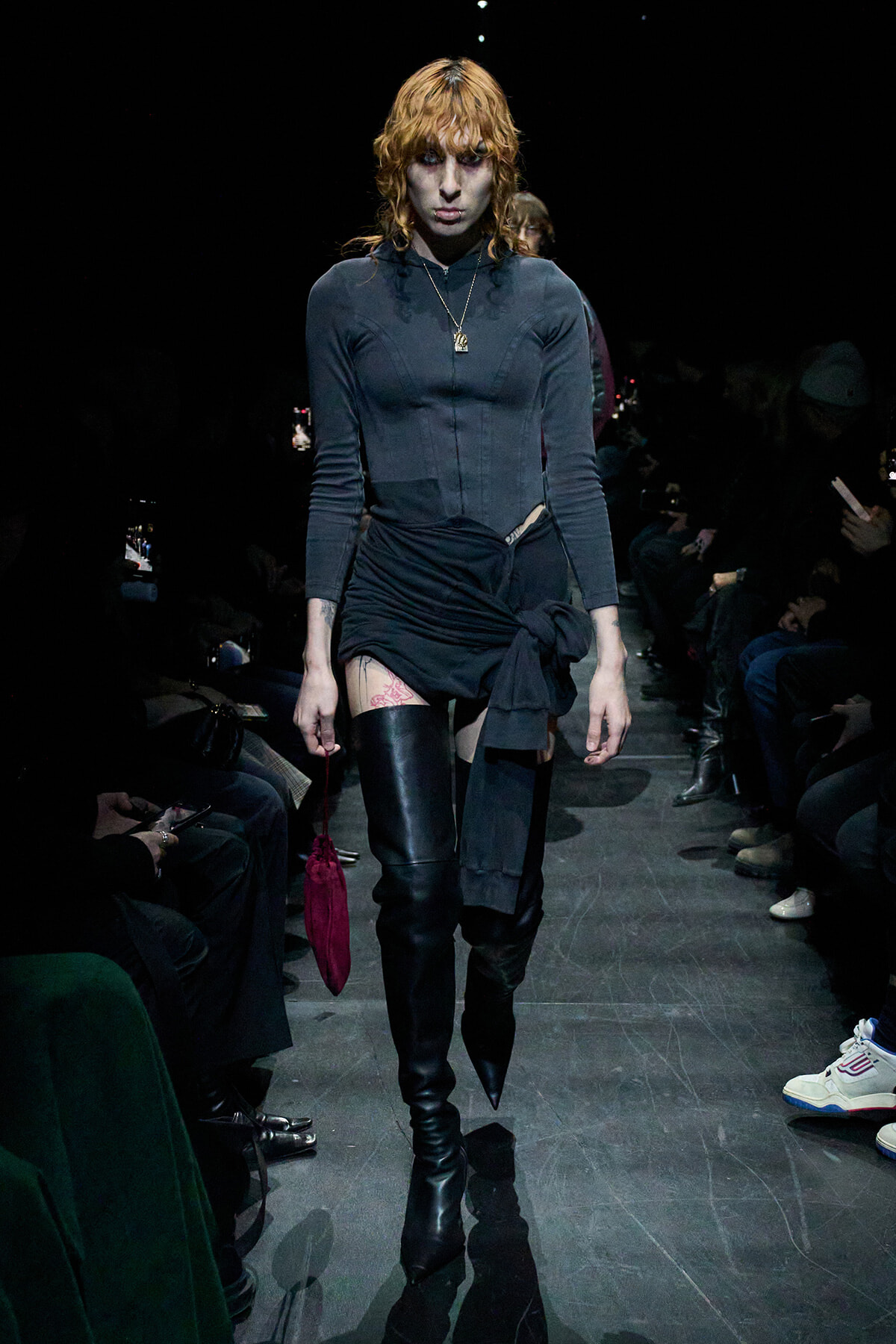 Model struts down a dark runway in a fitted dark grey top, black wrapped mini skirt, and tall black boots, with orange hair and a pendant necklace.