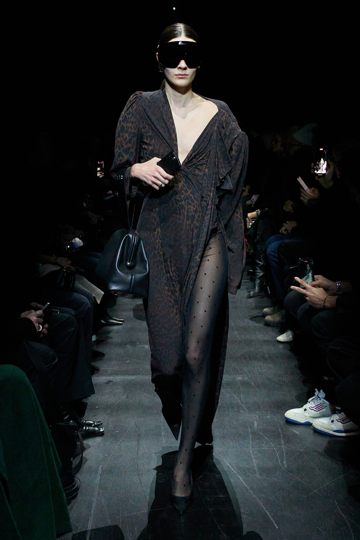 Model on a runway wearing a dark leopard-print wrap dress, oversized black sunglasses, and carrying a black bag with a phone in hand.