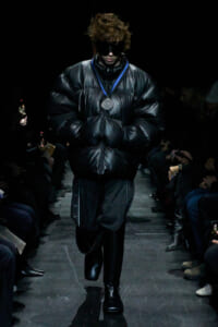 Model walks a runway in an oversized black puffer jacket, black boots, and dark sunglasses, with a silver medal on a blue ribbon around the neck.