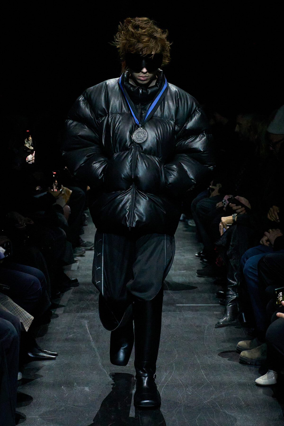 Model walks a runway in an oversized black puffer jacket, black boots, and dark sunglasses, with a silver medal on a blue ribbon around the neck.