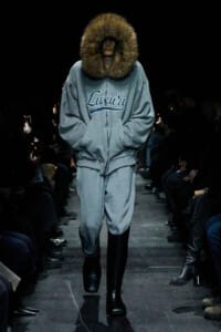 Runway model in a gray oversized hoodie with a large fur-trimmed hood and matching sweatpants, wearing black boots.
