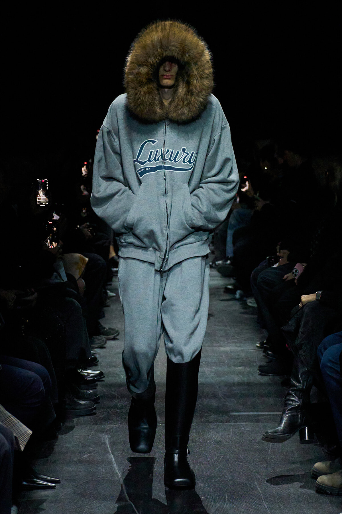 Runway model in a gray oversized hoodie with a large fur-trimmed hood and matching sweatpants, wearing black boots.