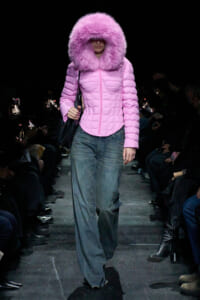 Model walks the runway in a pink padded coat with a large fluffy hood and blue jeans, carrying a black bag.