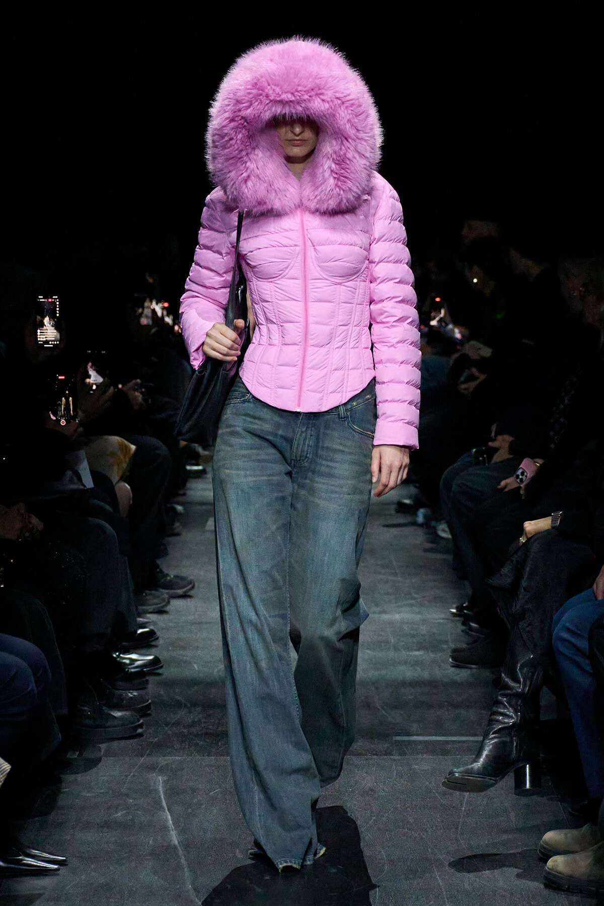Model walks the runway in a pink padded coat with a large fluffy hood and blue jeans, carrying a black bag.