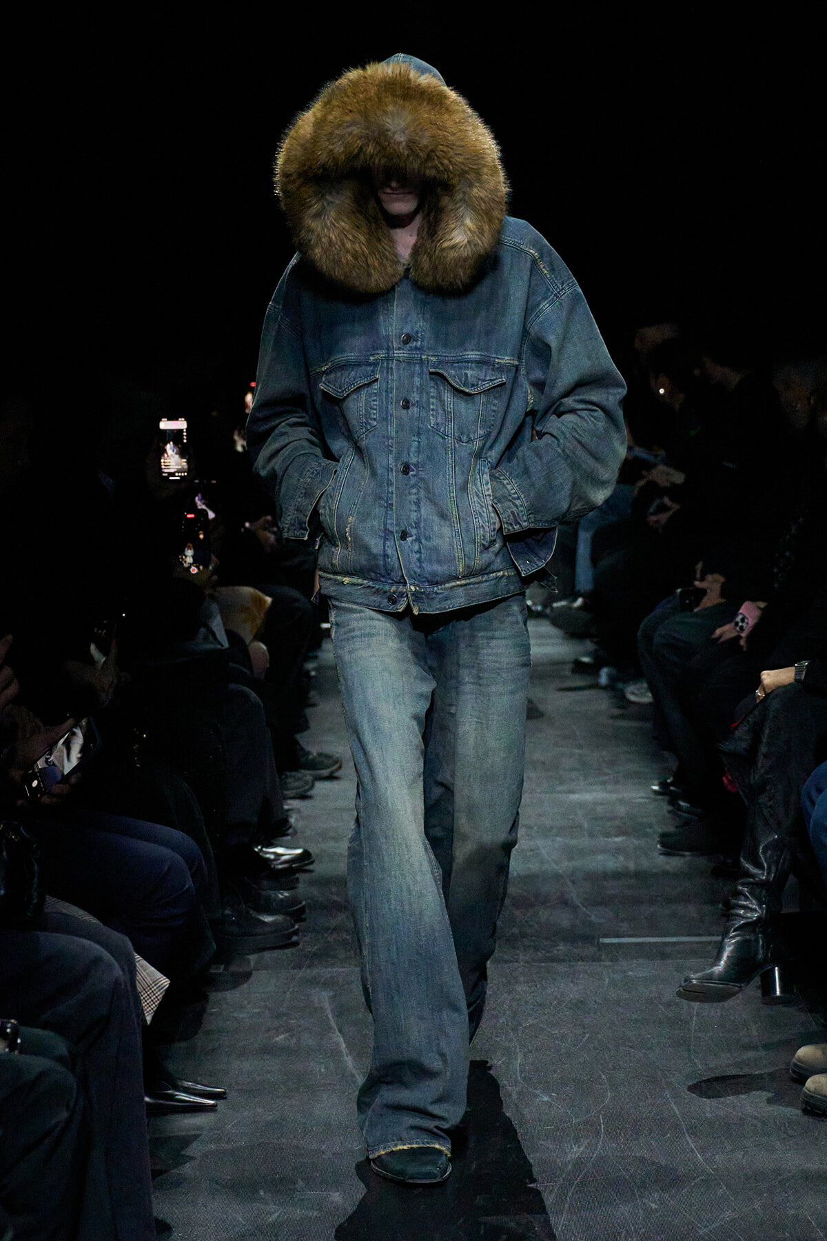 Model walks a fashion runway wearing an oversized blue denim jacket with a large fur-trimmed hood, hands in pockets, paired with faded jeans; dark audience lining the catwalk on both sides.