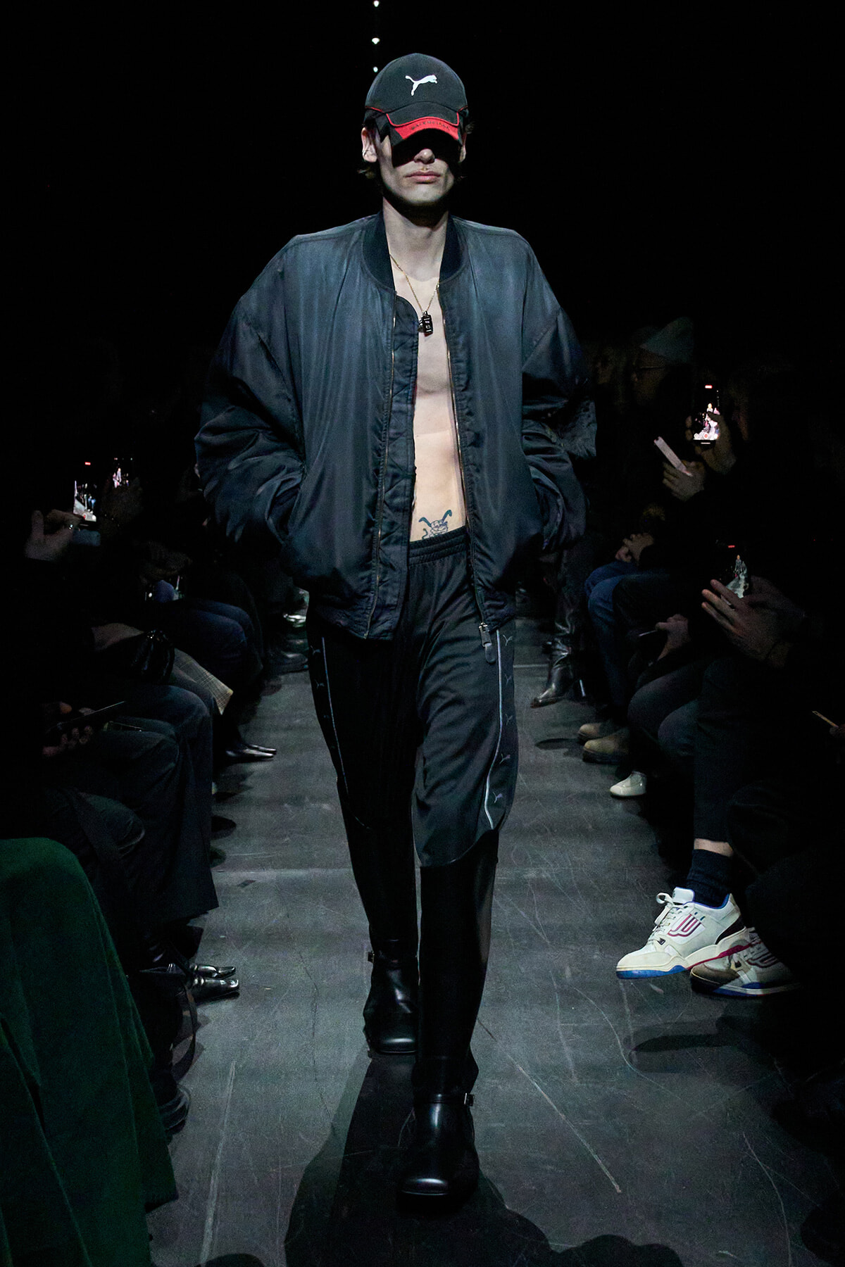 Male model walks the runway in a black bomber jacket open over a bare chest, wearing a black cap with a white logo and black boots; audience on sides.