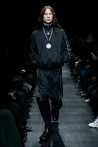 Male model walking a fashion runway in a black tracksuit with silver medal necklace; long dark hair and black boots.