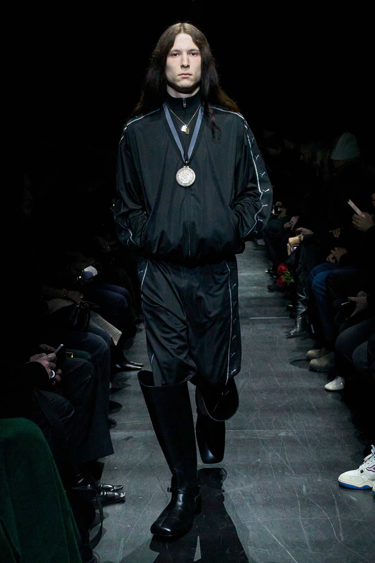 Male model walking a fashion runway in a black tracksuit with silver medal necklace; long dark hair and black boots.