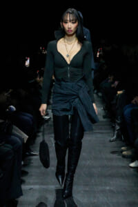 Model walking a dark runway in a black zip-front top, tight waist, layered jacket tied around the hips, and thigh-high boots.