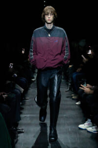 Male model walking a fashion runway in a burgundy and blue windbreaker and black leather pants, wearing glasses.