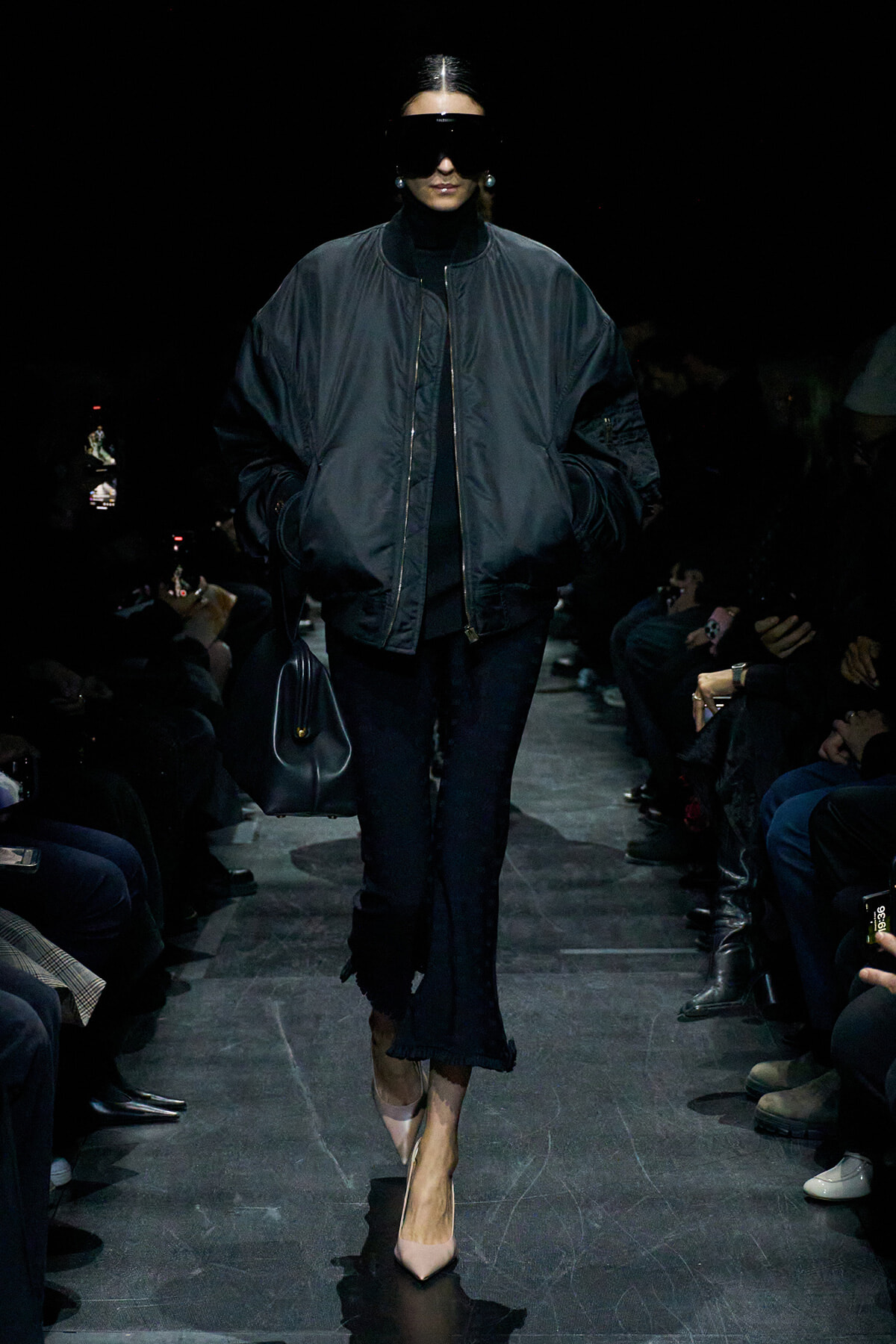 Model walks the runway in a oversized black bomber jacket, black turtleneck, dark jeans, nude pointed-toe heels, and a black handbag, wearing oversized sunglasses.
