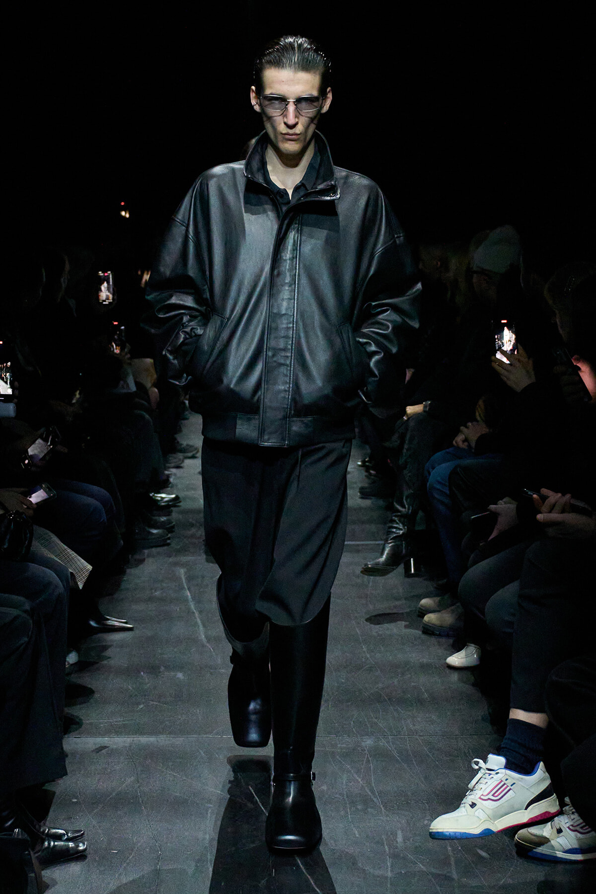 Male model on a dim runway wearing an oversized black leather jacket and dark pants, audience filming with phones nearby.