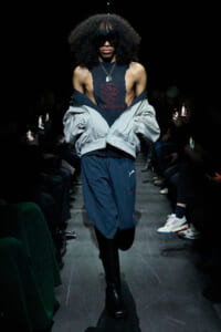 Model walks the runway in a black sleeveless top with red crest, a light gray jacket draped off the shoulders, and navy track pants with black boots.