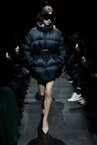 Model on a dark runway wearing an oversized black puffer coat belted at the waist, with futuristic sunglasses, walking toward the camera.