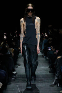 Model walks the runway in a sleeveless black top with cutouts, black leather pants, chunky boots, and a large black visor, showcasing tattoos on both arms amid a dark audience backdrop.