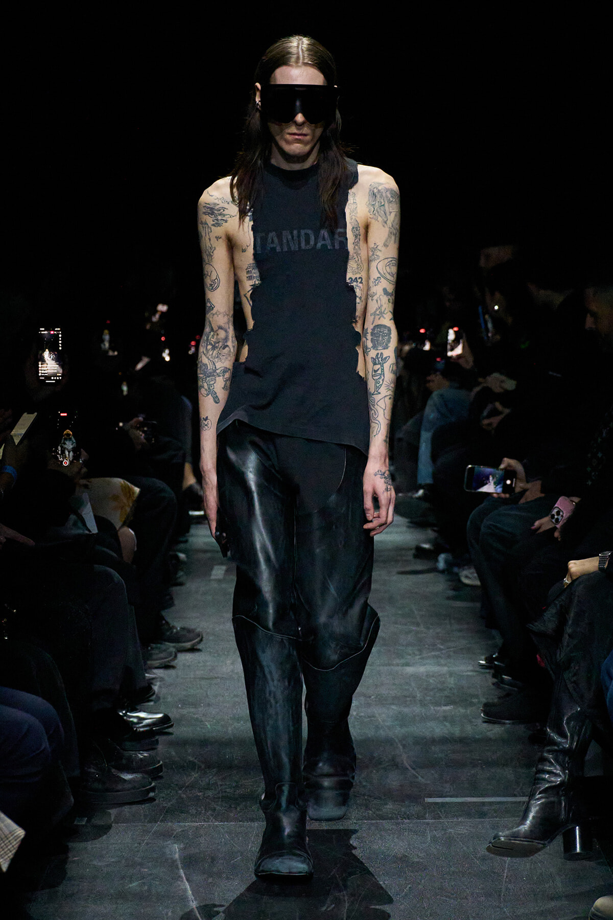 Model walks the runway in a sleeveless black top with cutouts, black leather pants, chunky boots, and a large black visor, showcasing tattoos on both arms amid a dark audience backdrop.
