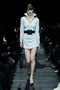 Model walking a runway in a white shirt-dress with an oversized black belt and pointed black heels, carrying a small black handbag.