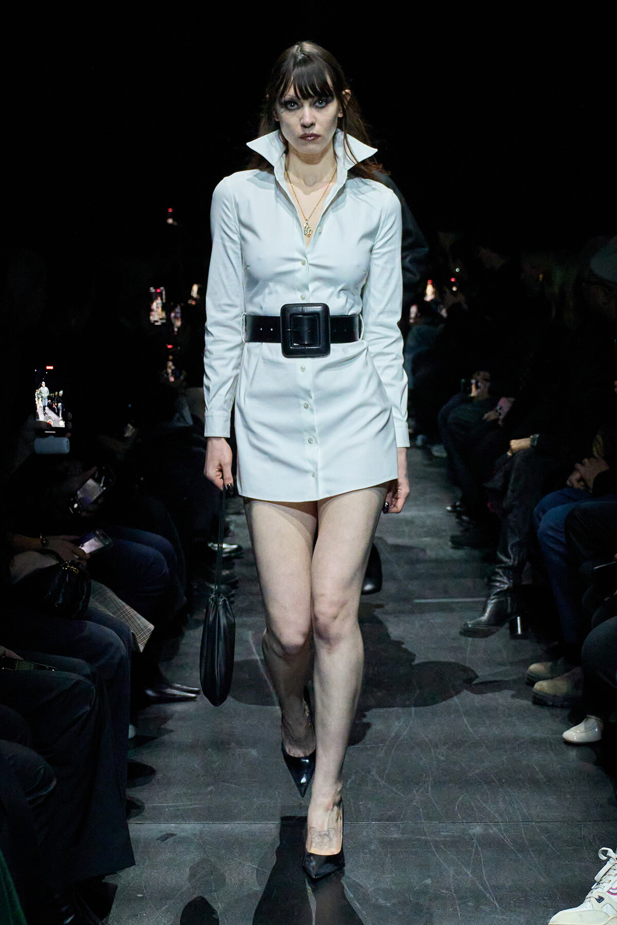 Model walking a runway in a white shirt-dress with an oversized black belt and pointed black heels, carrying a small black handbag.