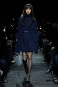 Model walks a dim runway in a navy knit turtleneck dress, cinched at the waist with a slim belt, wearing black heels and carrying a light blue bag.