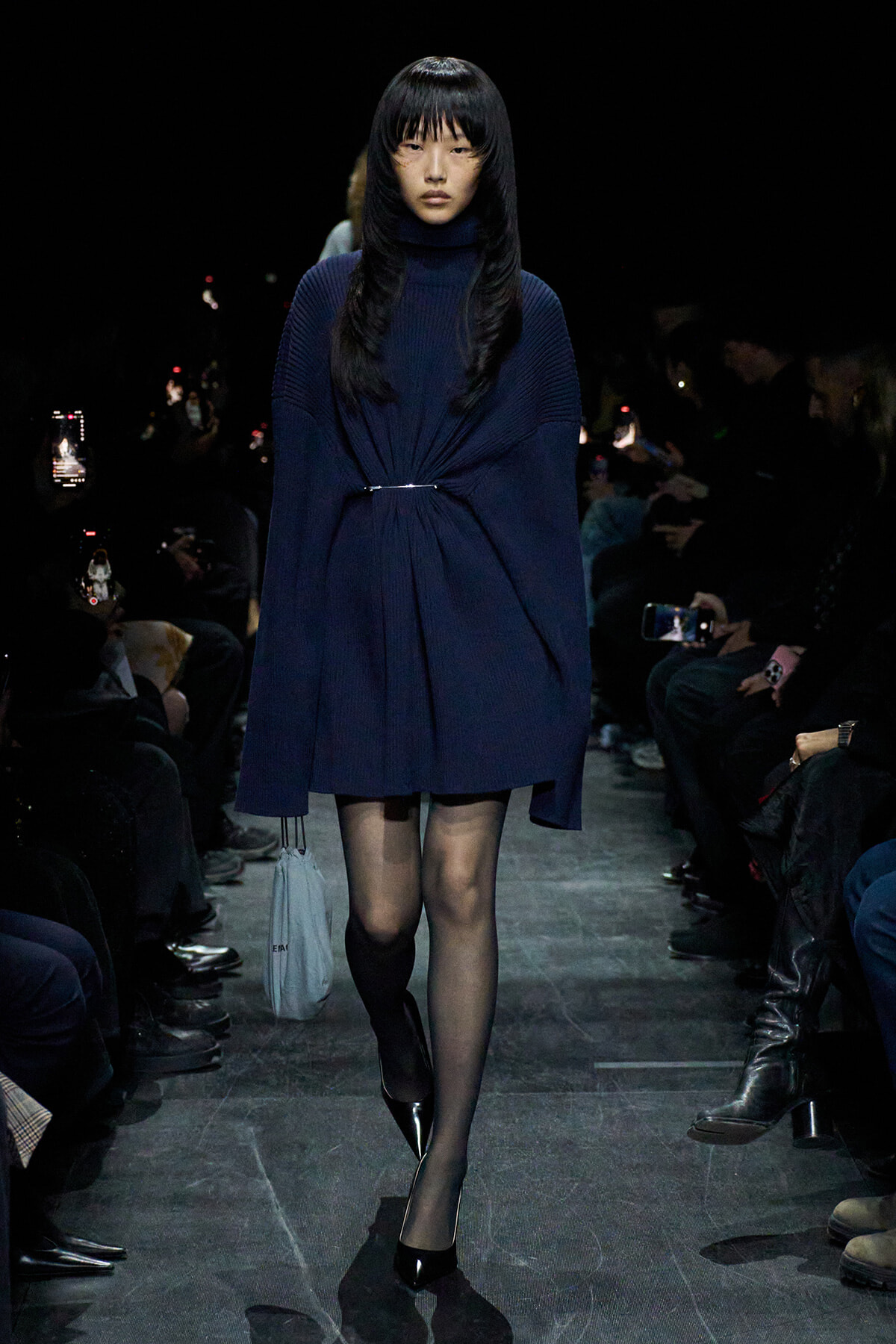 Model walks a dim runway in a navy knit turtleneck dress, cinched at the waist with a slim belt, wearing black heels and carrying a light blue bag.