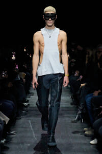 Male runway model wearing a light-gray sleeveless top, black pants, and oversized black goggles, walking down a fashion show runway.