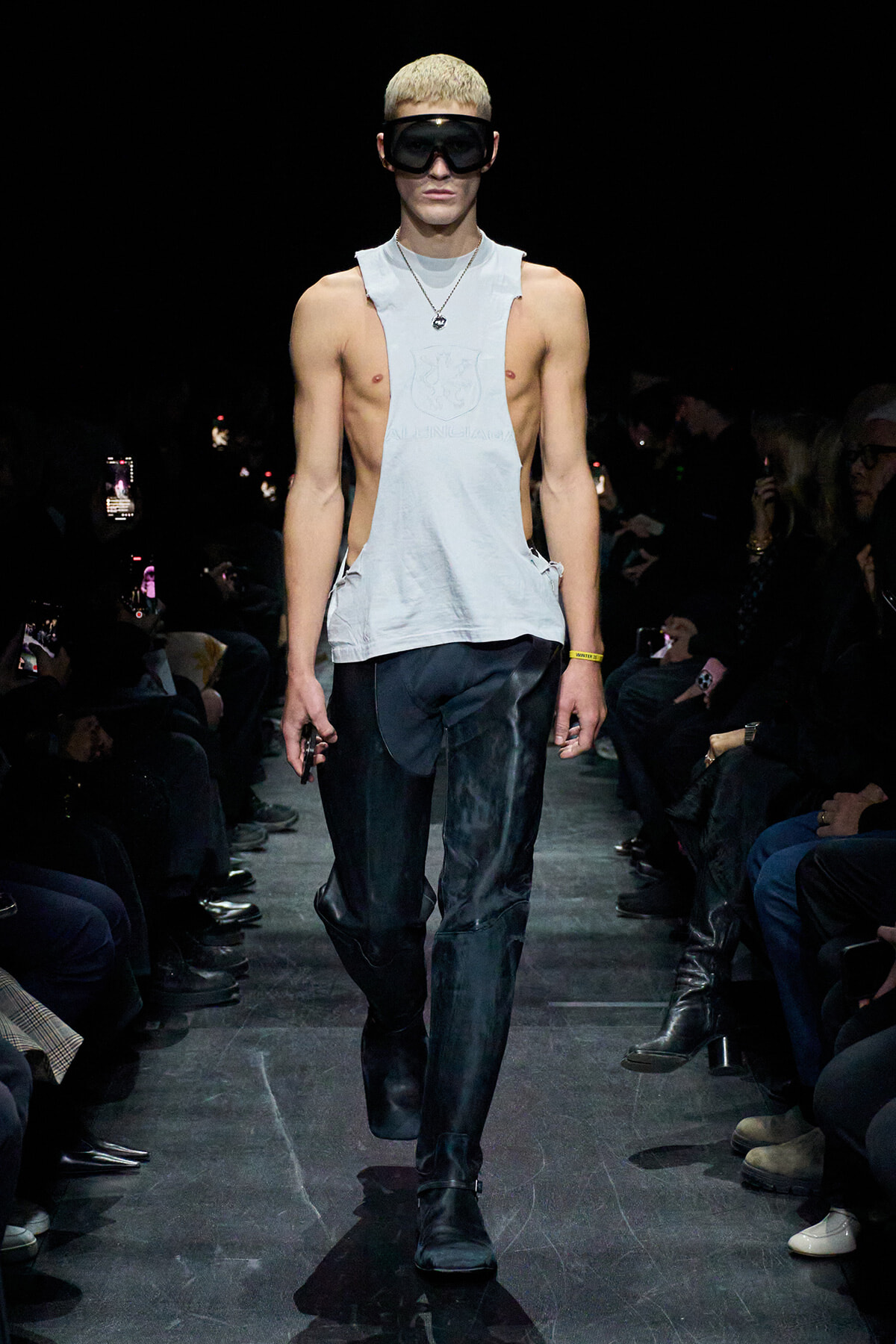 Male runway model wearing a light-gray sleeveless top, black pants, and oversized black goggles, walking down a fashion show runway.
