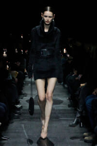 Model walks a dim runway in a black fur coat with a wide belt and nude heels, carrying a small black bag.