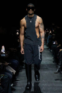Male model on a fashion runway wearing a ripped sleeveless black top, black track pants with vertical white lettering, chunky black boots, and oversized visor sunglasses.