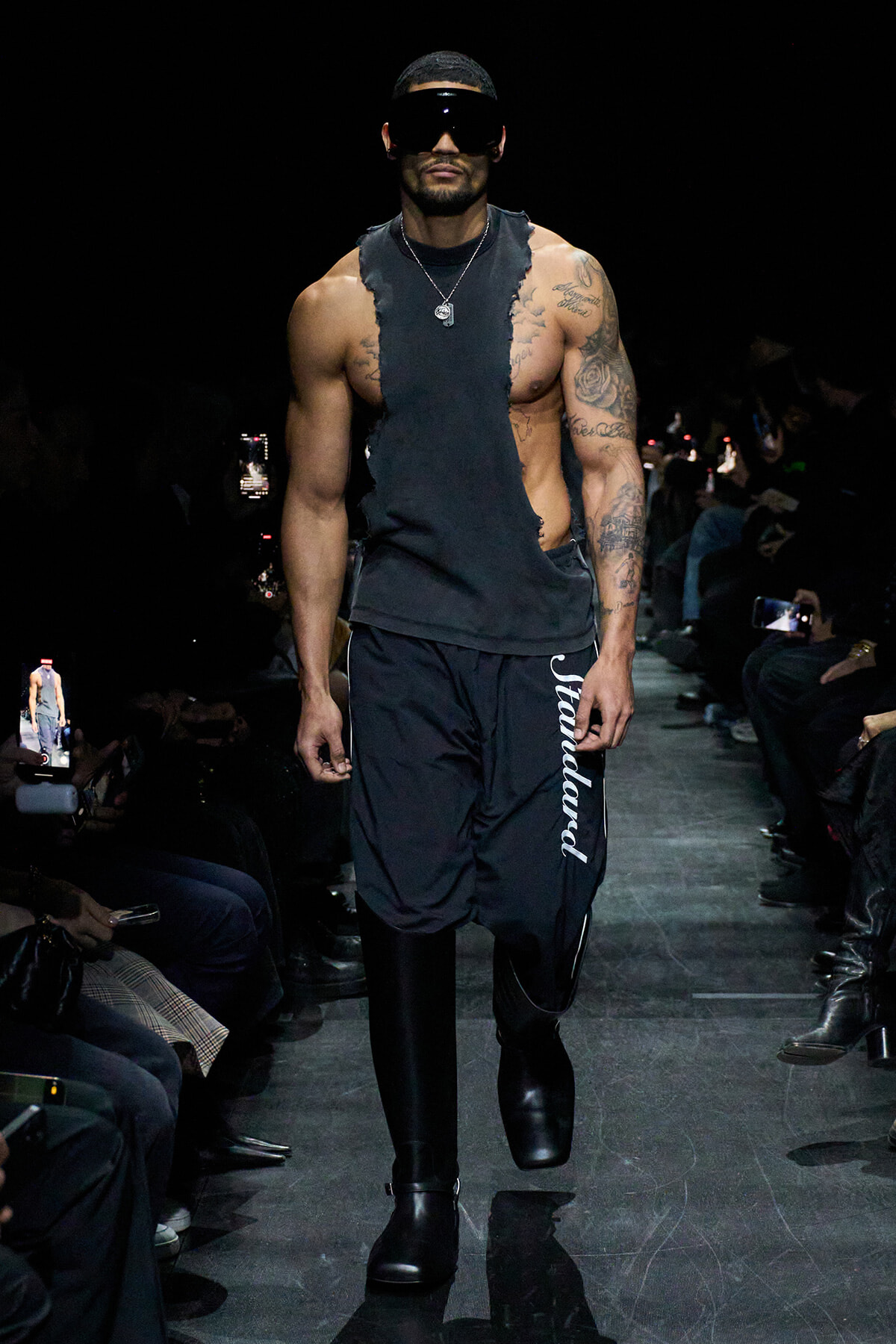 Male model on a fashion runway wearing a ripped sleeveless black top, black track pants with vertical white lettering, chunky black boots, and oversized visor sunglasses.