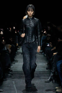 Model walking a dark runway in a black leather corset top and relaxed black pants.