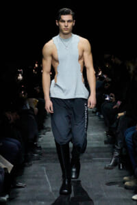Male model walking a runway in a light gray torn sleeveless top, black jogger pants, and tall black boots; silver pendant necklace visible; dark backstage lighting with audience on both sides.