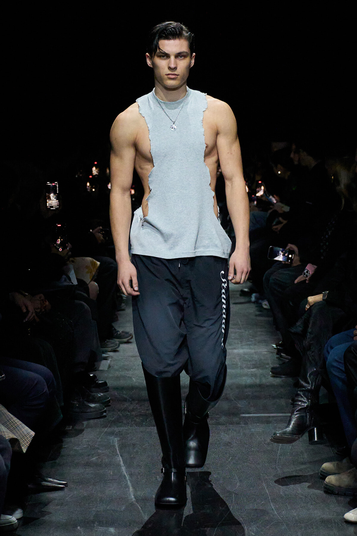 Male model walking a runway in a light gray torn sleeveless top, black jogger pants, and tall black boots; silver pendant necklace visible; dark backstage lighting with audience on both sides.