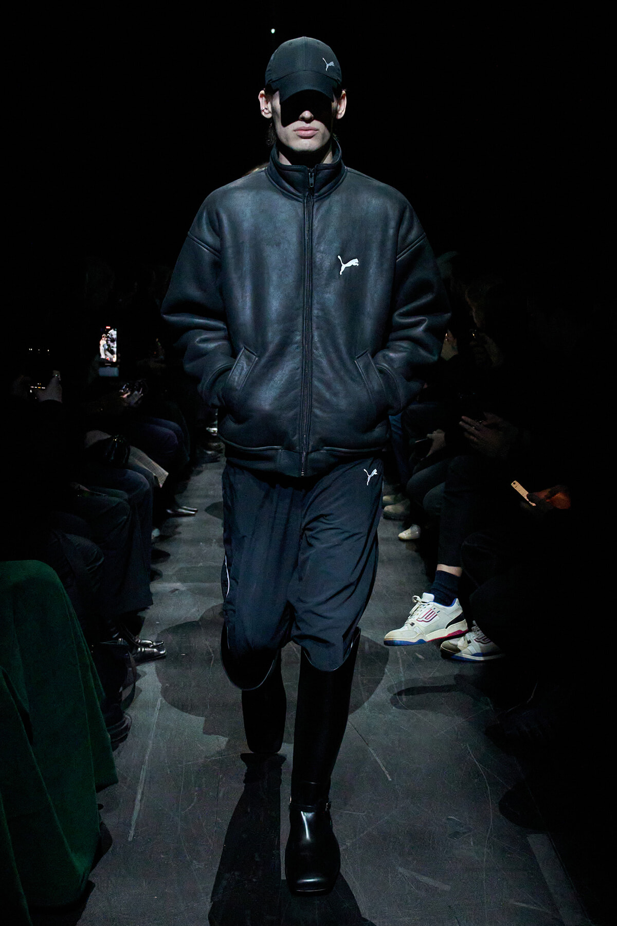 Model walking a dimly lit runway in all-black athleisure: black zip-up jacket with Puma logo, cap, and dark pants.