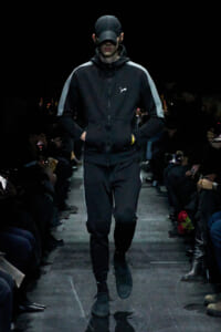 Male model walking a dark fashion runway in a black zip-up tracksuit with gray side stripes and a small white logo, wearing a black cap and hands in pockets.