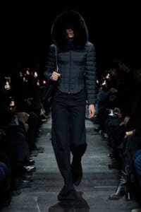 Model walks a dark runway in a quilted puffer with oversized fur hood; audience snapshots glow from the sides toward her.