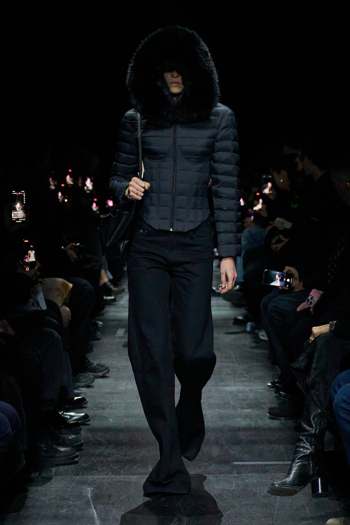 Model walks a dark runway in a quilted puffer with oversized fur hood; audience snapshots glow from the sides toward her.