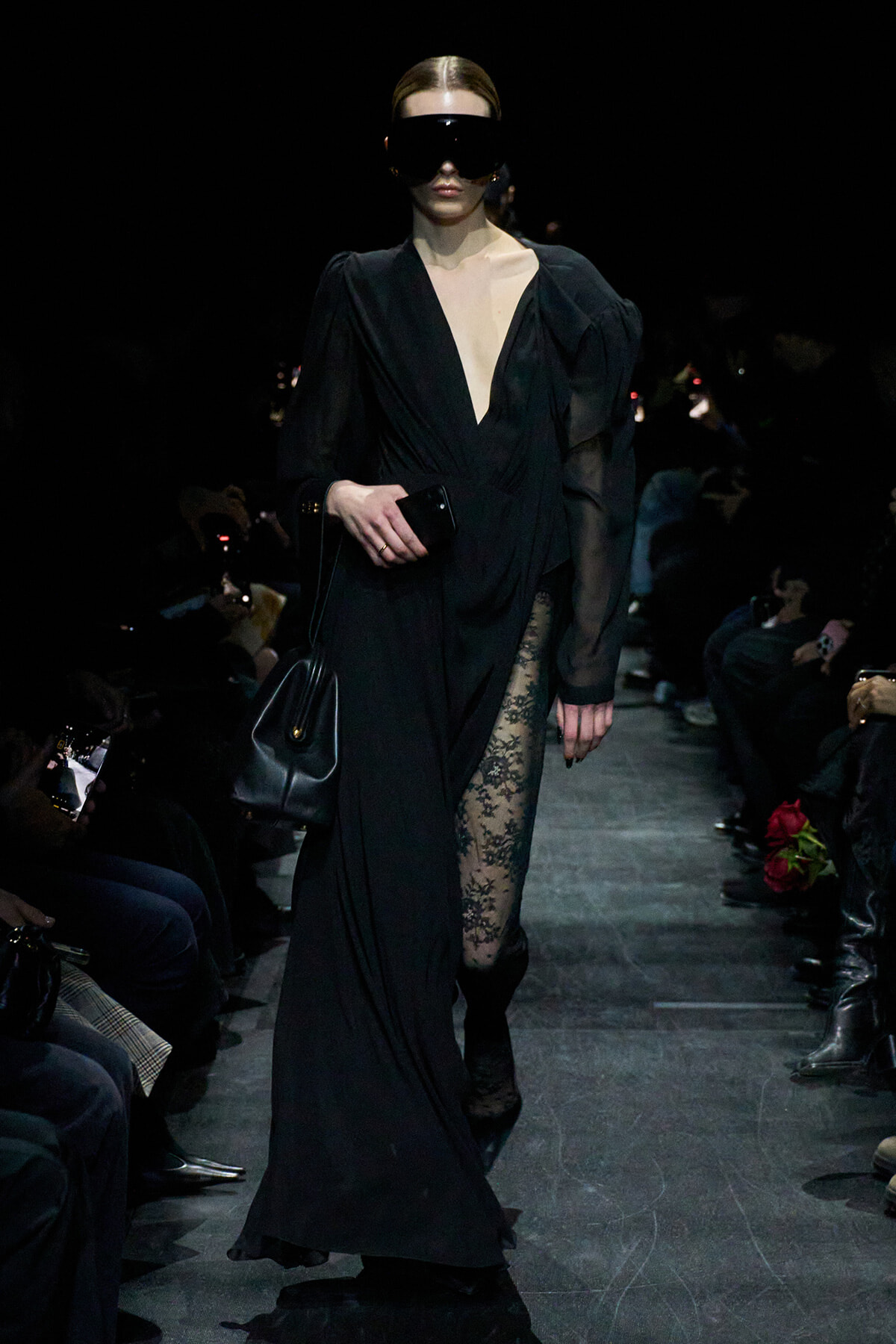 Model on a runway in a black gown with a plunging V-neck, sheer sleeves, and a lace panel at the leg, wearing a large black mask and carrying a clutch.