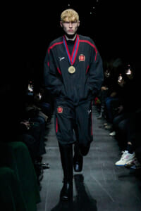 Male model walking a runway in a black tracksuit with red stripes, wearing a gold medal necklace.