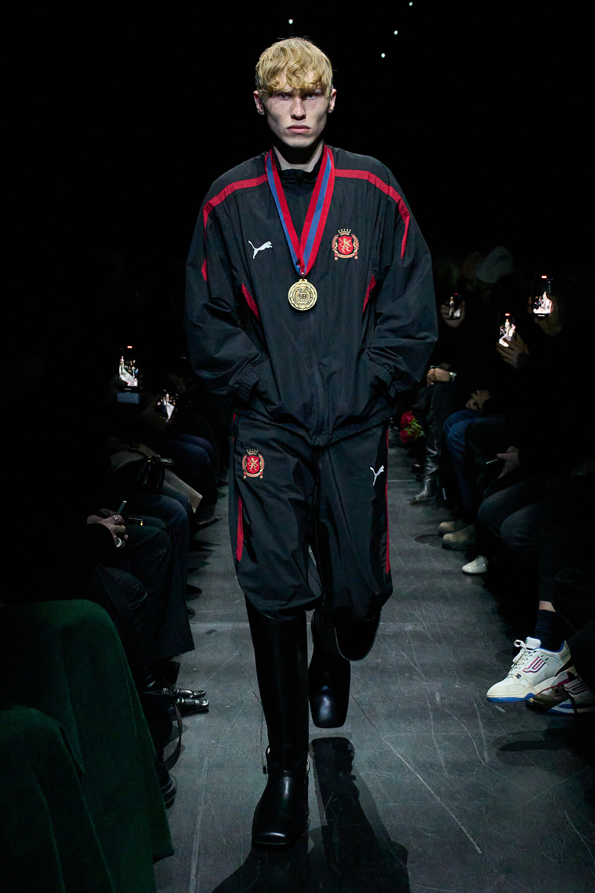 Male model walking a runway in a black tracksuit with red stripes, wearing a gold medal necklace.