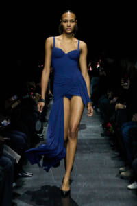 Model walks a fashion-show runway in a cobalt blue, form-fitting dress with a ruched bodice and flowing wrap skirt, black heels.