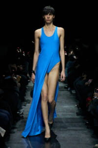 Model on a runway wearing a bright blue sleeveless dress with a high slit, black heels, and dark audience seating along the sides.