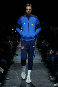 Model walking a fashion runway in a blue Puma tracksuit with crest, hands in pockets, wearing glasses and white sneakers.