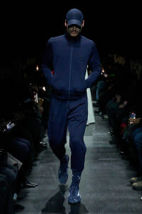 Male model wearing a navy tracksuit and cap walks a dim fashion runway, hands in pockets, focused forward.